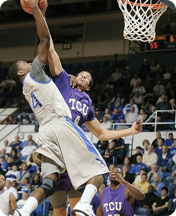Basketball player in white jersey jumping for a dunk with defender in purple attempting to block. Energetic action in packed indoor gym.