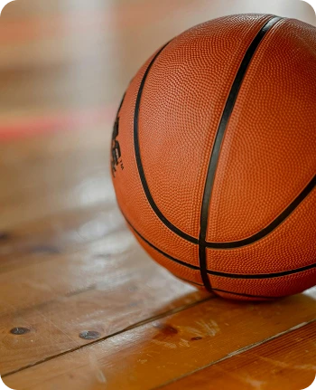 Close-up of a basketball resting on a polished wooden gym floor. The lighting highlights the orange texture, evoking a sense of anticipation and competition.